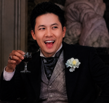 A joyful man in a formal black suit, patterned grey vest, and white flower boutonniere holds a glass of red wine in what appears to be a period drama setting.