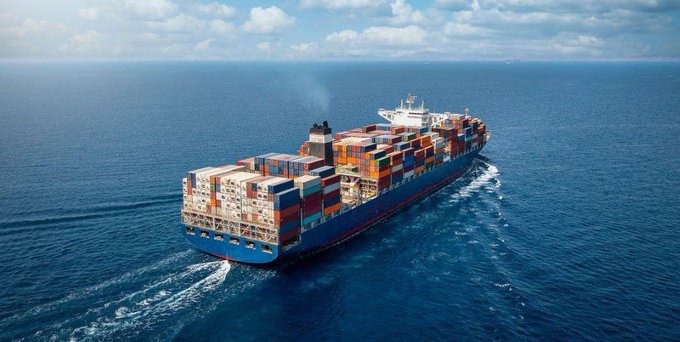 An aerial photograph of a large blue container ship loaded with colorful shipping containers sailing across the open ocean under a partly cloudy sky.
