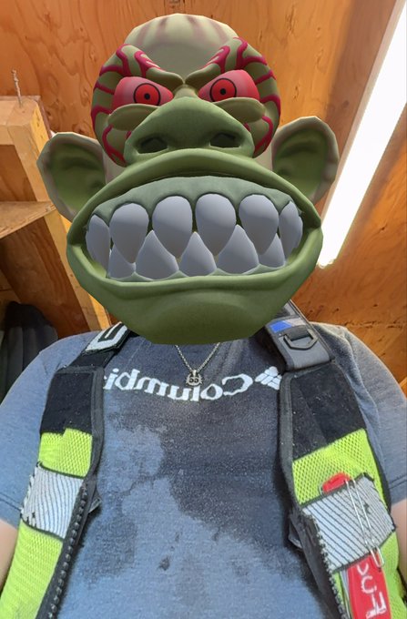 A person wears a Columbia t-shirt with a reflective safety vest and a pendant necklace, their face replaced with a cartoonish green troll mask filter.