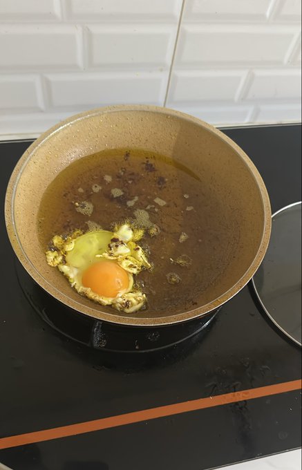 A single egg sizzles in oil within a well-used brown frying pan on a black stovetop, capturing a candid "cooking time" moment as the white begins to set around the bright yolk.