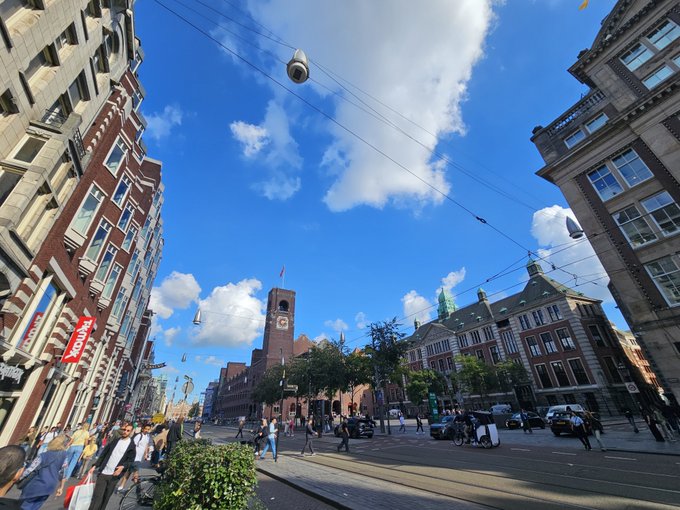 A bright, sunny day in a European city street, likely Amsterdam, featuring classic buildings, pedestrians, and tram cables crisscrossing the blue sky with fluffy clouds.
