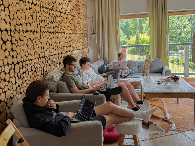 Four young men relaxing on couches in a cozy living room with a distinctive log-wall feature, each working on their laptops.