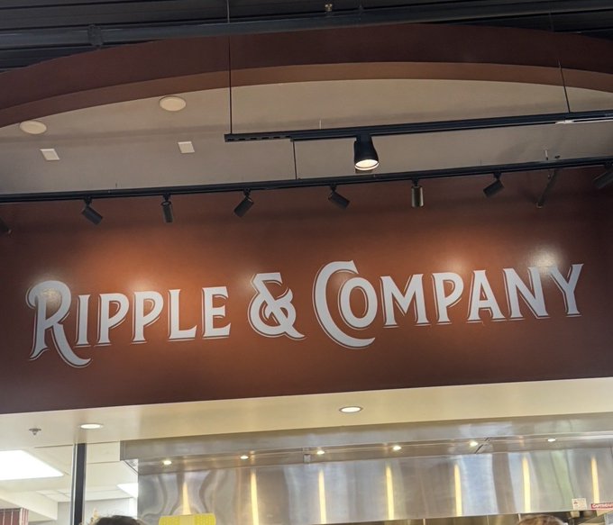 The image shows a restaurant with an overhead sign that reads "Ripple & Company", indicating its name and possibly the type of food served there. The sign is hanging from the ceiling, making it easily visible for customers. In front of the restaurant, there are people present, suggesting that the establishment is open for business.