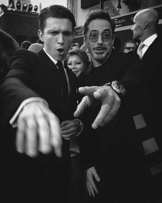 A black-and-white selfie taken at a movie premiere showing Tom Holland and Robert Downey Jr. pointing forward toward the camera.