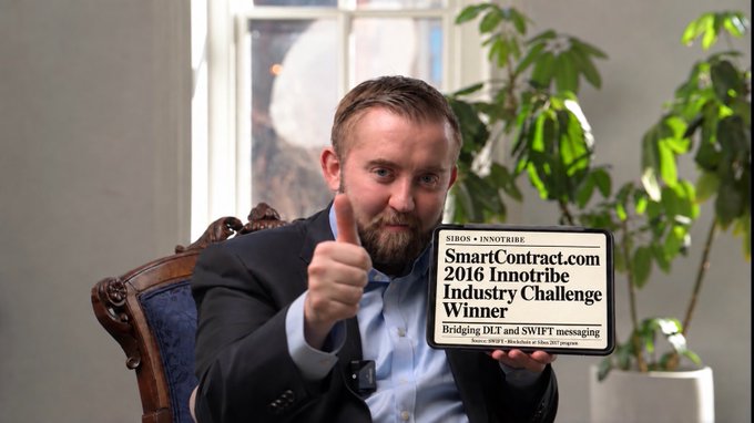 A bearded man in a suit gives a thumbs-up while holding a tablet that reads 'SmartContract.com 2016 Innotribe Industry Challenge Winner'.