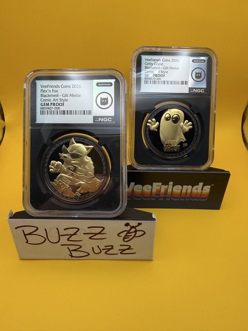 Two graded VeeFriends coins in NGC slabs displayed against a yellow background, labeled 'FLEX'n Fox' and 'Crusty Ghost' (Blackened - Gilt Medal), with a handwritten sign reading 'BUZZ BUZZ'.