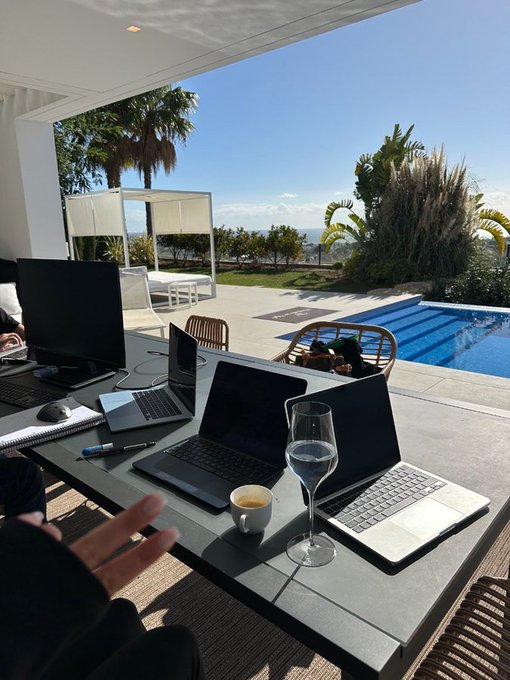 An outdoor workspace with two laptops, a desktop monitor, a wine glass, and an espresso cup overlooks a swimming pool and tropical landscape.