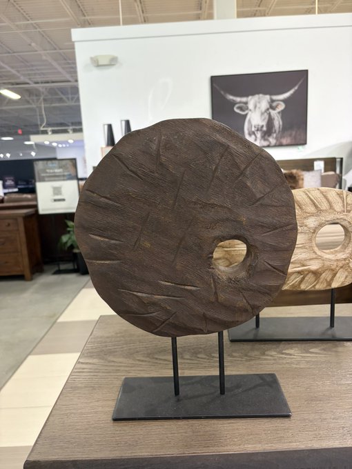 A large, ancient-looking Rai stone coin sculpture is displayed on a black metal stand in a furniture store, with a bull artwork visible in the background. The post notes the shopper’s accidental framing of the bull behind the coin, adding humor to the image.