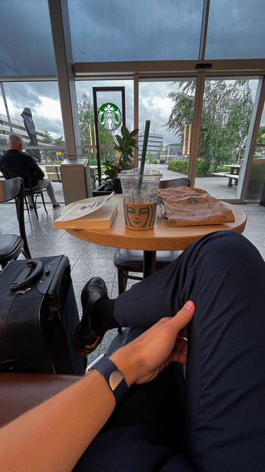 A first-person view shows a person relaxing at a Starbucks with an iced drink, wearing dark pants and black loafers, with a suitcase beside them near a large window.