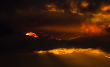 A dramatic sunset scene with the sun partially obscured behind dark, fiery clouds, casting golden crepuscular rays across a deep orange and black sky.
