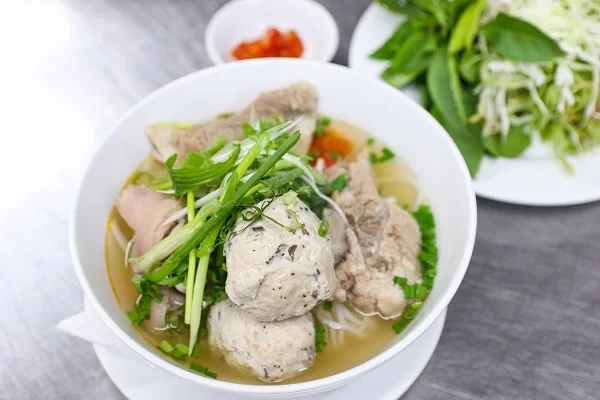 A bowl of Vietnamese pho or pork noodle soup with meatballs, sliced pork, and fresh herbs, served alongside a plate of greens and bean sprouts.
