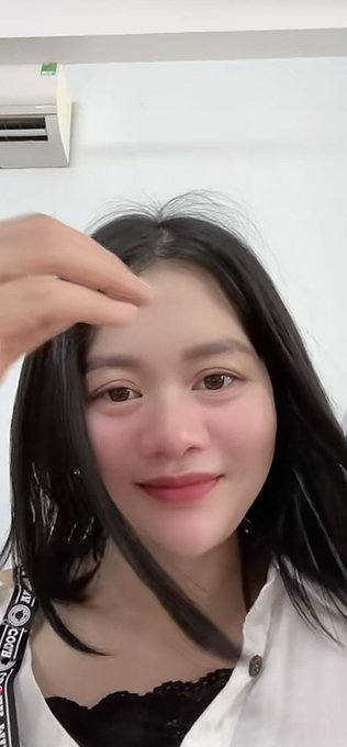 A young woman with dark hair takes a close-up selfie, wearing a white shirt with a black lace-trimmed top underneath, touching her hair near her forehead.