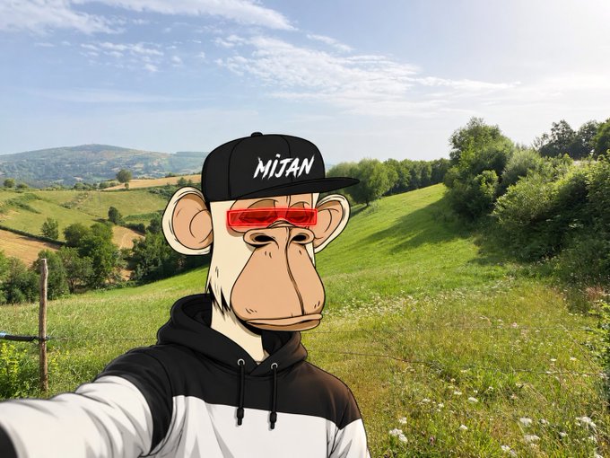 A selfie of a person wearing a Bored Ape-style monkey mask with red shutter sunglasses and a black 'MITAN' cap, set against a scenic background of rolling green hills and blue sky.