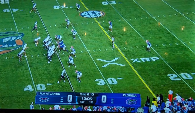A football game between Florida Atlantic Owls and Florida Gators shows a tied 0-0 score with 12:09 remaining in the 1st quarter, while an "XRP" logo is prominently displayed on the field near the 30-yard line. The XRP branding on the field ties directly into the post's promotion of XRP cryptocurrency, suggesting a sponsorship deal or marketing tie-in during the broadcast.