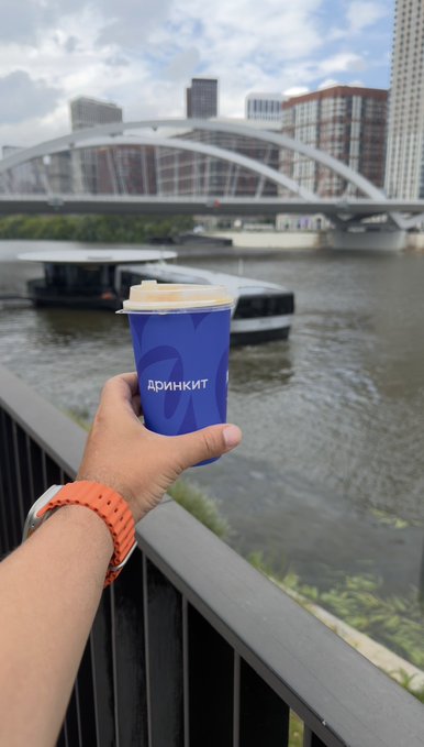 A hand holds a blue coffee cup with the text 'АРИАНКИТ' (Ariankit) in front of a river, bridge, and city skyline, with an orange watch band visible on the wrist.
