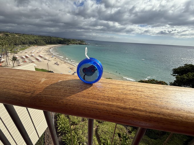 A small blue circular charm or pendant featuring a cartoon animal character sitting on a wooden balcony railing, overlooking a scenic beach with turquoise water and umbrellas.