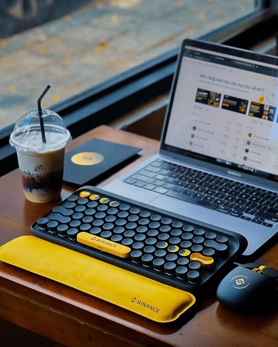 A Binance-branded keyboard and wrist rest sit beside a laptop and iced coffee, capturing a productive café work session that embodies the "work hard, play hard" lifestyle.