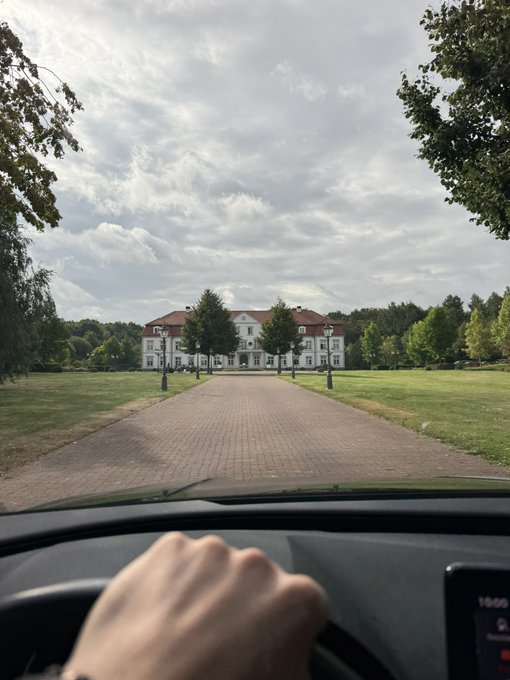 A first-person view from inside a car driving on a brick pathway toward a large white historic building or estate surrounded by green lawns and trees under a cloudy sky.