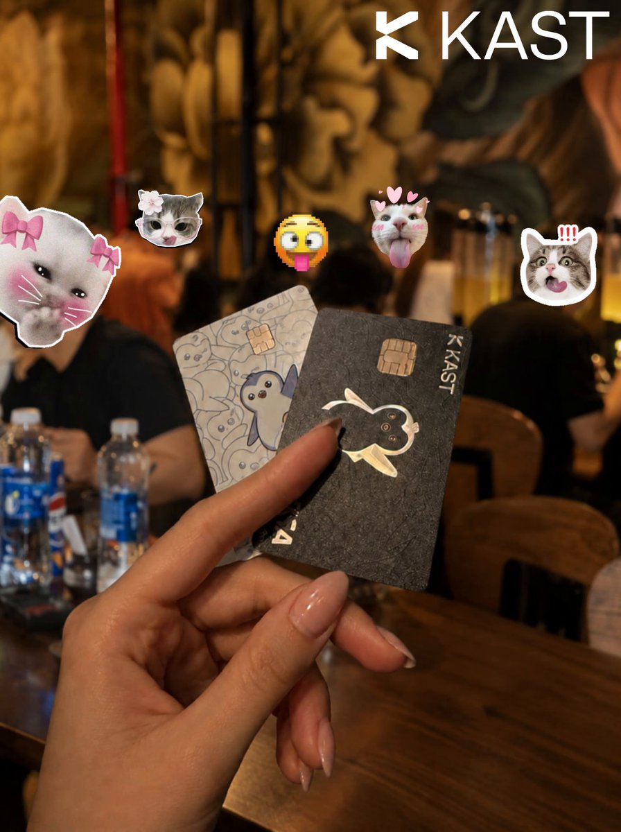 The image features a hand holding two credit cards, one of which has a cat sticker on it. The other card appears to be a KAST credit card. A person's nails are painted with a light brown color, adding a personal touch to the scene. In the background, there is another person sitting at a table with a bottle and a cup placed in front of them.