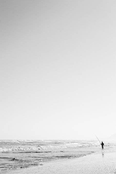 A minimalist black-and-white photograph of a solitary figure standing in the shallow surf of a vast ocean, holding a fishing rod, emphasizing the vast, empty sky and shoreline.