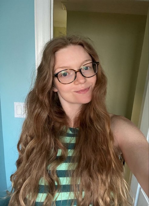 A young woman with long, wavy reddish-brown hair and tortoiseshell glasses smiles softly for a selfie, her hair's natural volume and flow playfully matching her caption about waking up with hair that still "mogs."