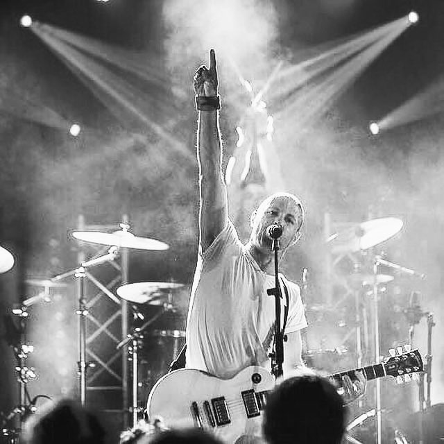 A black-and-white photo of a bald musician performing on stage, raising his fist while singing into a microphone and holding a Les Paul guitar, surrounded by stage lights and a drum kit.