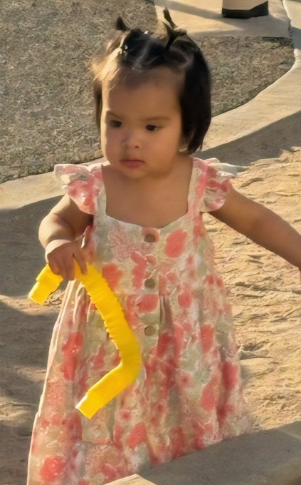 The image features a young girl wearing a pink floral dress, holding a yellow object that appears to be a toy or a piece of art. The girl seems to be walking outdoors with her hands in her pockets. In the background, there are some trees visible, adding to the outdoor setting. The photo is related to a post on social media where someone has captioned it: "The best thing in life is not about if I'm rich or poor.. It is about spending time with my 1.3 months old granddaughter.. She makes me cried today by calling me 'Ong' Vietnamese for grandpa."