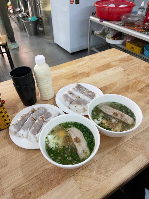 A Vietnamese meal on a wooden table featuring two bowls of banh cuon (steamed rice rolls) in soup and two plates of additional rice rolls, accompanied by a bottle of soy milk.