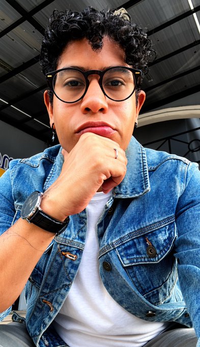 A close-up selfie of a young man with curly dark hair and glasses, wearing a denim jacket over a white t-shirt, resting his chin on his hand.