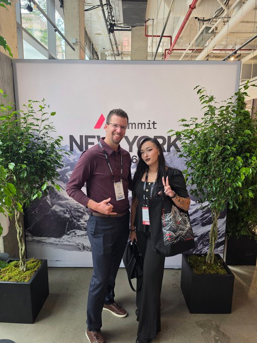 Two people pose in front of a "Summit New York" banner, with a man in a maroon polo shirt and glasses standing next to a woman in black attire who is holding up a peace sign. They are flanked by potted plants and the woman carries a tote bag.