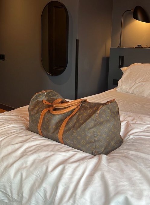 A brown Louis Vuitton monogram duffel bag rests on a white bed in a dimly lit room with a modern lamp and oval mirror in the background.