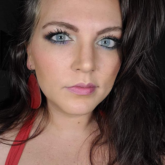 A close-up portrait of a woman with long dark hair, striking blue eyes with colorful eyeshadow, pink lipstick, and large red leather earrings.
