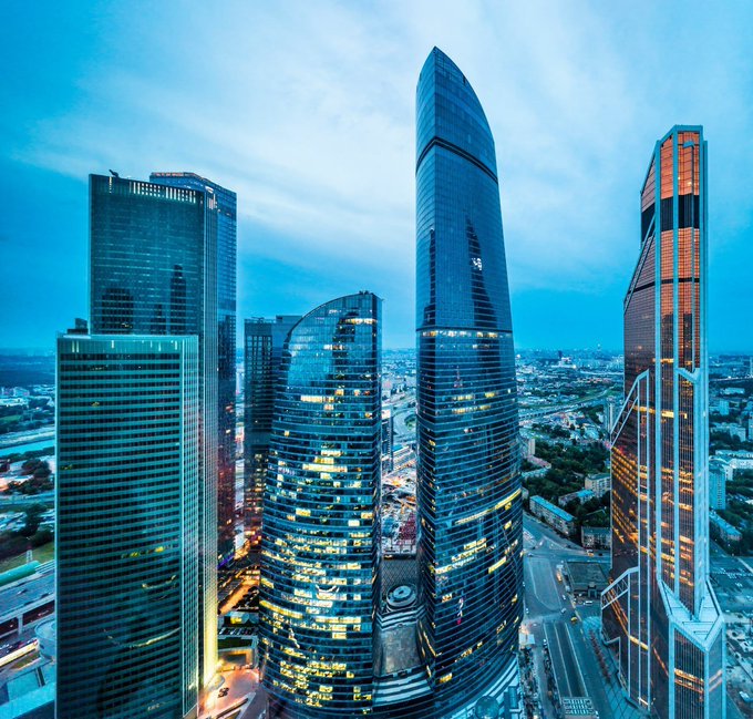 A twilight aerial view of a modern city skyline featuring several tall skyscrapers with reflective glass facades, including a prominent tower with a distinctive curved, asymmetrical shape.