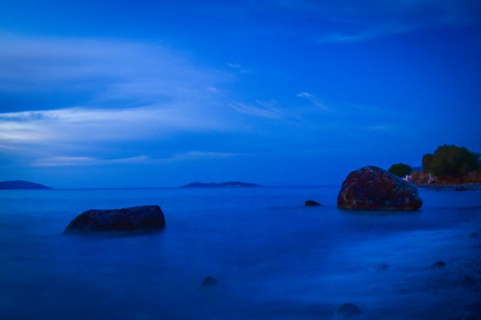 A serene twilight seascape featuring calm waters, scattered rocks in the foreground, and a deep blue cloudy sky with distant land masses on the horizon.