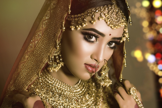 A glamorous portrait of a woman adorned in elaborate gold bridal jewelry, including a maang tikka, heavy necklace, and large nose ring, with a red bindi on her forehead.