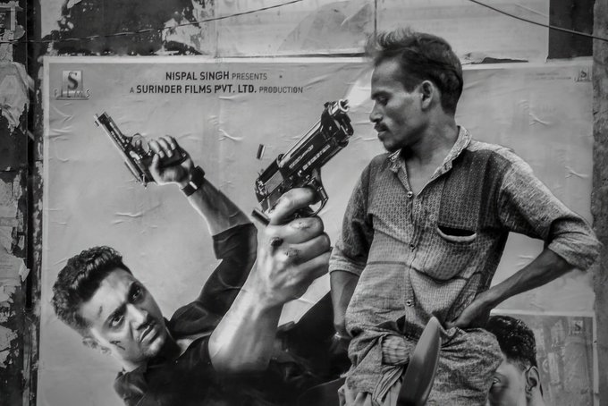 In this black and white photo, two men are standing next to each other with guns drawn, possibly posing for a picture or participating in an action scene. The man on the left has his gun pointed towards the right side of the image, while the man on the right holds his gun pointed towards the left side. Both men have their hands on their hips and appear to be engaged in conversation or discussion. 

The background features a poster with text that reads "NFT SOLD নায়ক নিলবন দিজনিলবন দিজনিলবন দিজনিলবন দিজনিলবন দিজনিলবন দিজনিলবন দিজনিলবন দিজনিলবন দিজনিলবন".