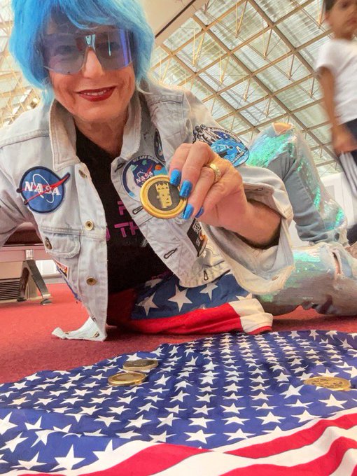 Woman in a NASA jacket, blue wig, and star-spangled outfit holding up a Bitcoin-themed pin while sitting on an American flag at what appears to be a convention.