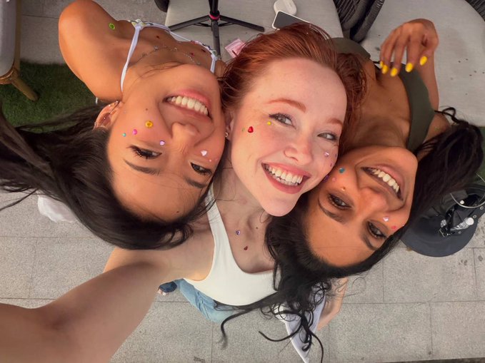 An overhead selfie of three young women lying down close together, all smiling brightly with colorful jewel stickers decorating their faces.