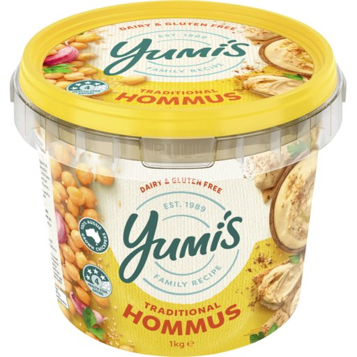 A 1kg tub of Yumi's Traditional Hommus, a dairy and gluten-free product established in 1989, featuring a yellow lid and images of chickpeas and herbs on the label.