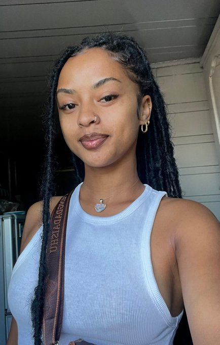A young woman with long braided hair, a nose ring, and gold earrings takes a selfie wearing a light blue tank top with a patterned strap.