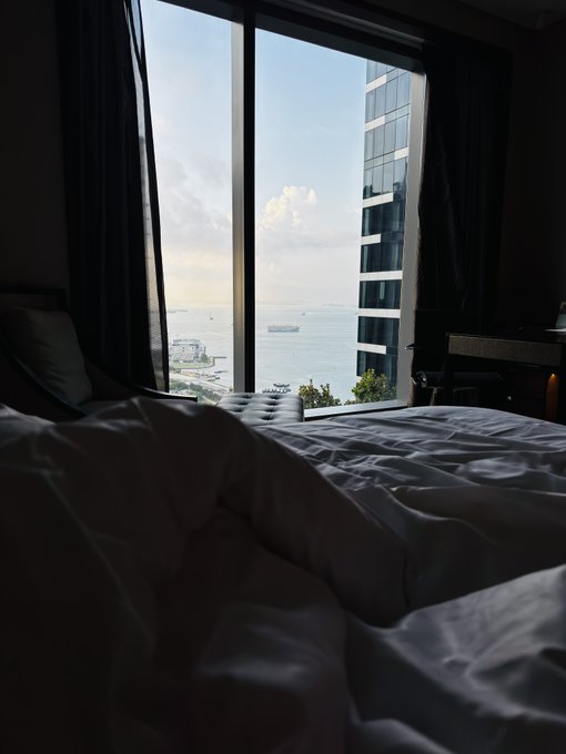Morning sunlight streams through a hotel window, illuminating rumpled white sheets on an unmade bed while offering a serene view of the ocean and a neighboring high-rise under a partly cloudy sky—a perfect waking moment to greet the "good morning sky."