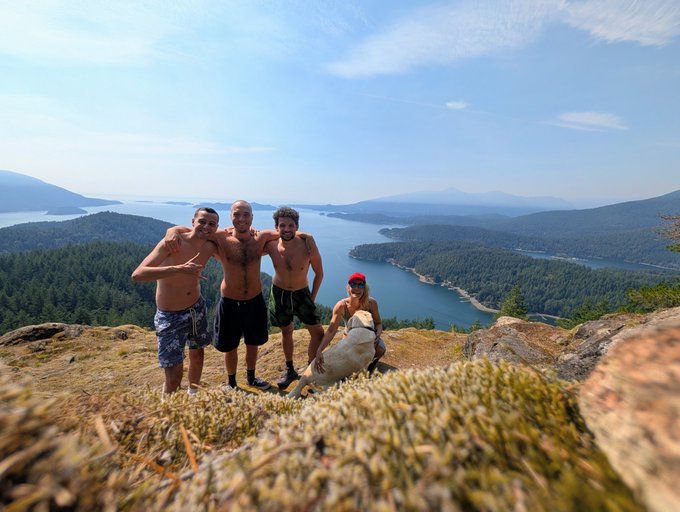 Four shirtless men pose triumphantly on a rocky mountain summit, with one seated holding what appears to be a small bear cub, against a stunning backdrop of forested islands and blue lakes under a clear sky. The image captures the adventurous spirit of the post's "build in the bear" theme, showing the group proudly displaying their unusual mountain-top encounter with wildlife.