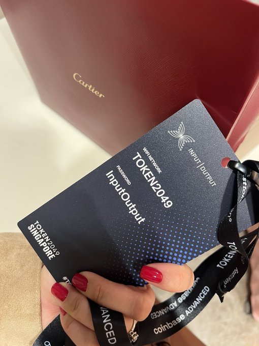 A person with red nail polish holds a black 'TOKEN2049 Singapore' attendee pass labeled 'InputOutput', with a red Cartier shopping bag visible in the background.