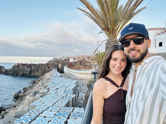 A selfie of a smiling couple—a woman in a brown top and a man wearing a blue cap and sunglasses—posing with a coastal view, palm tree, and decorative tiled steps behind them.