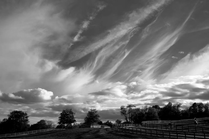 A black and white landscape photograph showing dramatic, sweeping cirrus clouds over a rural scene with silhouetted trees and a wooden fence.