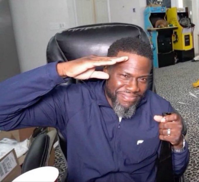 A man in a dark blue zip-up jacket sits in a black office chair, smiling and saluting with one hand while holding a cigar in the other, with arcade machines visible in the background—capturing a confident, victorious "gm" moment ready to seize the day.