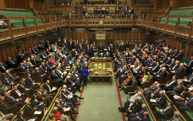 The image shows the interior of the House of Commons in the British Parliament, with MPs seated on green benches facing each other and the Speaker's Chair at the far end. This iconic chamber setting relates to the post, as it depicts the very body launching the inquiry into banks restricting cryptocurrency-linked companies, illustrating the parliamentary setting where this six-week consultation on financial de-banking practices will take place.