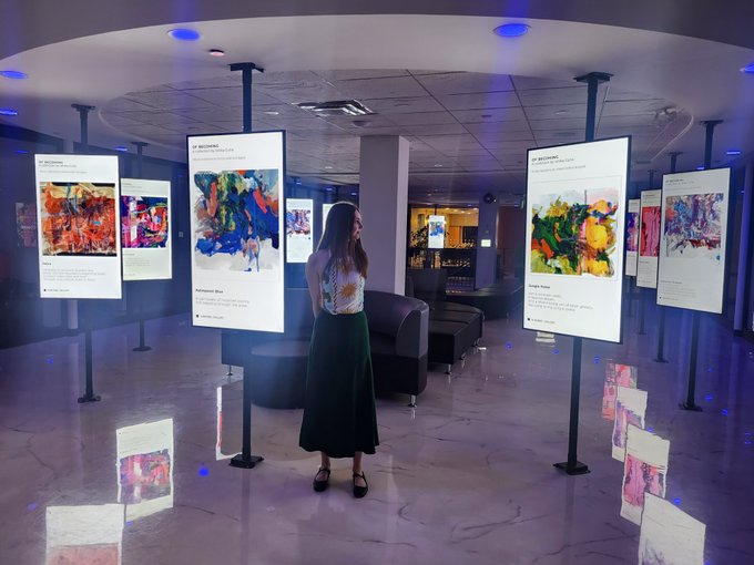 A woman in a green skirt stands in a modern exhibition space viewing digital artworks displayed on illuminated vertical screens, with her reflection visible on the polished floor.