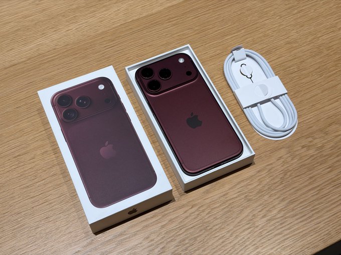 The image shows two burgundy/dark red iPhone 18 Pro Max boxes on a wooden surface, each displaying the phone's back with its distinctive triple-camera system and Apple logo, alongside a white charging cable. The post appears to be a giveaway promotion claiming to offer these iPhones to followers who like, retweet, and comment—though notably, Apple has not released an "iPhone 18 Pro Max" model, and no iPhone has ever been navy blue, suggesting this is a misleading scam post using staged product imagery.
