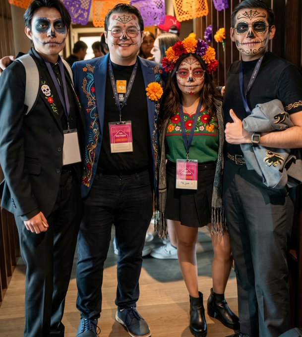 Four people pose together at what appears to be a Day of the Dead celebration, wearing sugar skull face paint, colorful embroidered clothing, and lanyards, with orange marigold decorations overhead.